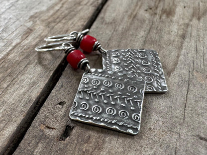 Dangle earrings featuring a diamond-shaped silver charm with embossed patterns, suspended from red African glass trade beads on silver ear wires.